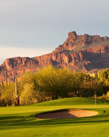 Golf course with green fairway and sand bunker, red rock mountain backdrop, desert vegetation and homes in foreground