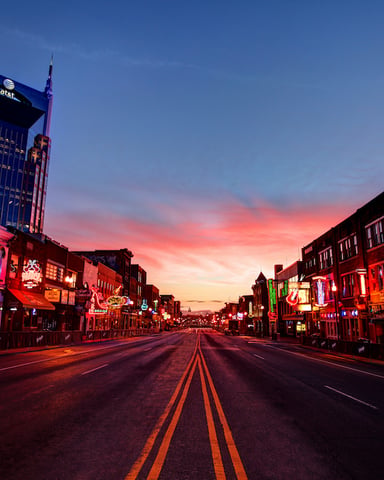 Broadway street at dusk with neon-lit storefronts, tall buildings, and colorful sunset sky