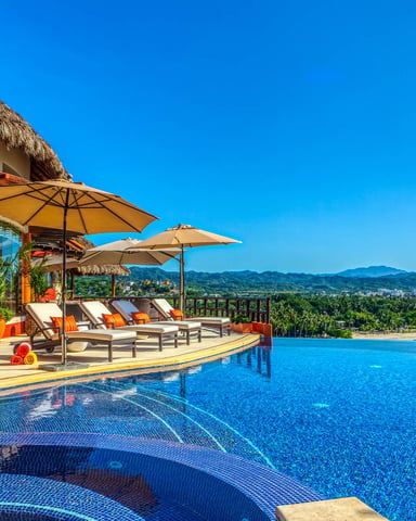 Infinity pool overlooking tropical coastline with thatched-roof villa, lounge chairs, umbrellas, and mountain views under clear blue sky