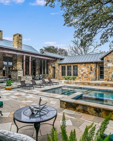 Stone house with metal roof overlooking patio, pool, fire pit, and lounge seating under large tree