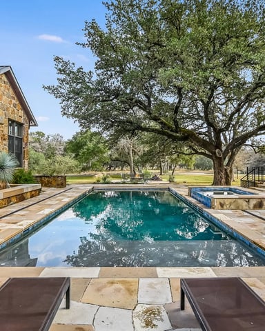Stone house with rectangular swimming pool, patio seating, and large oak tree overlooking open pasture