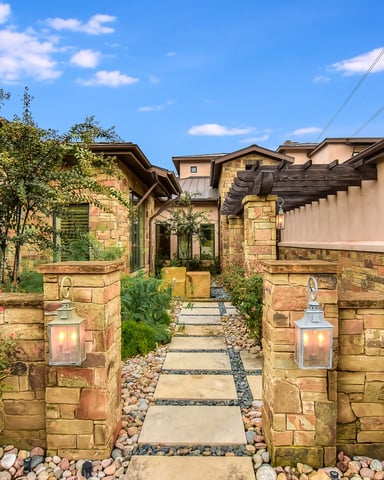 Stone courtyard pathway with flowering shrubs, lanterns on pillars, and modern stone houses with pergolas