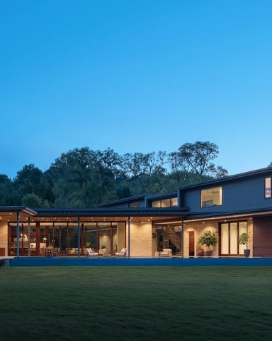 Modern single-story home with brick and metal exterior, extensive glass walls, and illuminated interior at dusk, surrounded by lawn and trees.