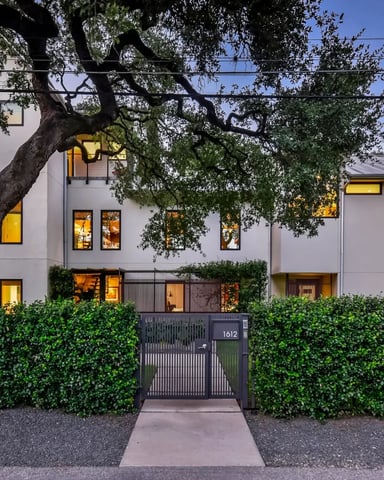 Modern white house with large oak tree, lit windows, and hedged front gate at dusk