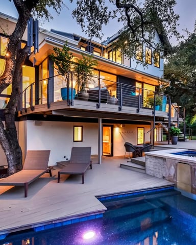 Modern two-story home with illuminated interior, backyard pool, lounge chairs, and mature trees at dusk