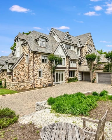 Stone and cream Tudor-style house with multiple gables overlooking a lake, paved driveway, manicured lawn, and landscaped gardens.