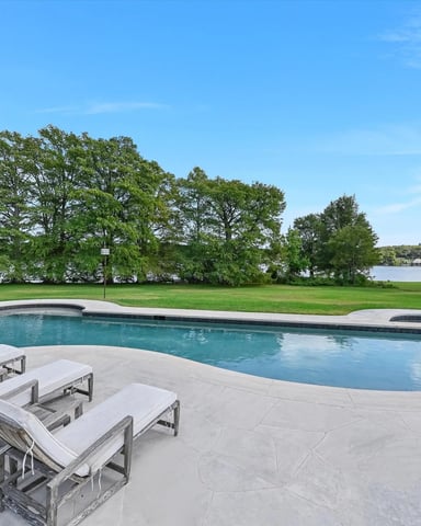 Swimming pool and spa with lounge chairs on patio, overlooking manicured lawn and waterfront with trees