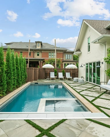 Backyard with rectangular swimming pool, lounge chairs, white house with green shutters, brick house, tall green hedges, and patio seating area