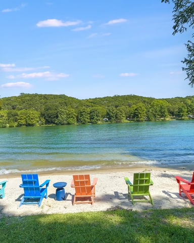 Sandy beach with colorful Adirondack chairs facing calm turquoise lake, forested hills beyond, docks and kayaks visible
