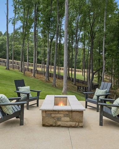 Outdoor fire pit seating area with Adirondack chairs on concrete patio, surrounded by tall trees and manicured lawn with fencing in background