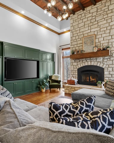 Living room with gray sectional sofa, green built-in cabinetry with TV, stone fireplace with wood mantel, wood beams, and large windows with wood frames.