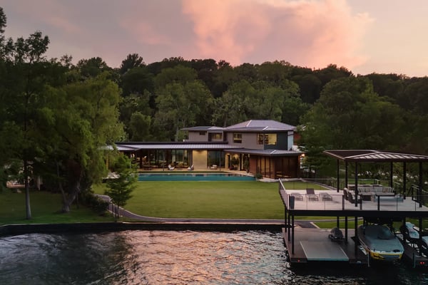 Modern waterfront home with pool, manicured lawn, and boat dock at dusk surrounded by trees