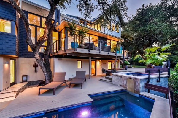 Modern two-story home with illuminated interior, backyard pool, lounge chairs, and mature trees at dusk