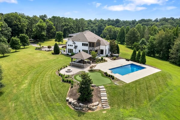 Modern white house with pool, patio, and putting green on expansive manicured lawn surrounded by trees