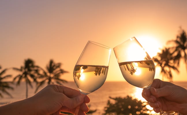 Two hands clinking wine glasses at sunset with palm trees and ocean in background