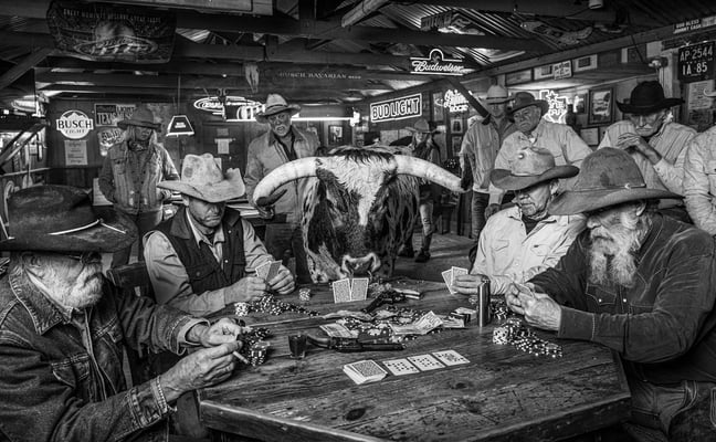 Black and white photo of cowboys playing poker around a table with a longhorn steer in a rustic bar decorated with beer signs
