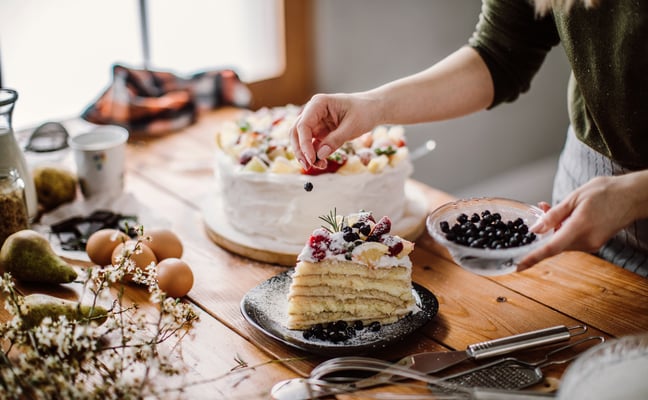 Person decorating layered cake with berries and powdered sugar on wooden table with fresh ingredients
