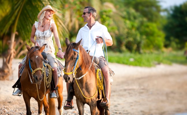 Couple horseback riding on a sandy beach path lined with palm trees