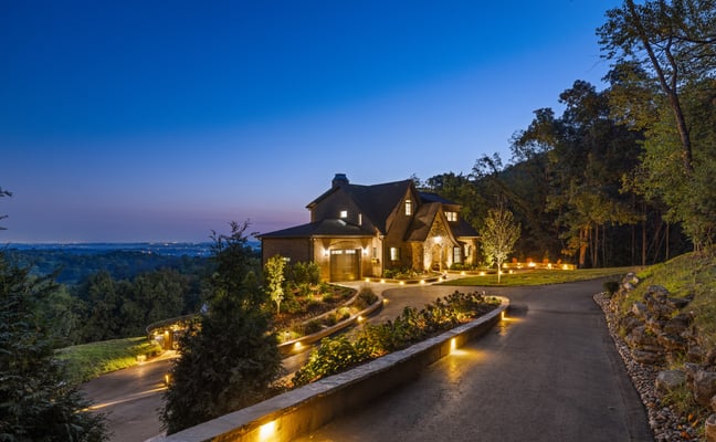 Illuminated brick house on hillside at dusk with curved driveway, landscaping, and valley view beyond