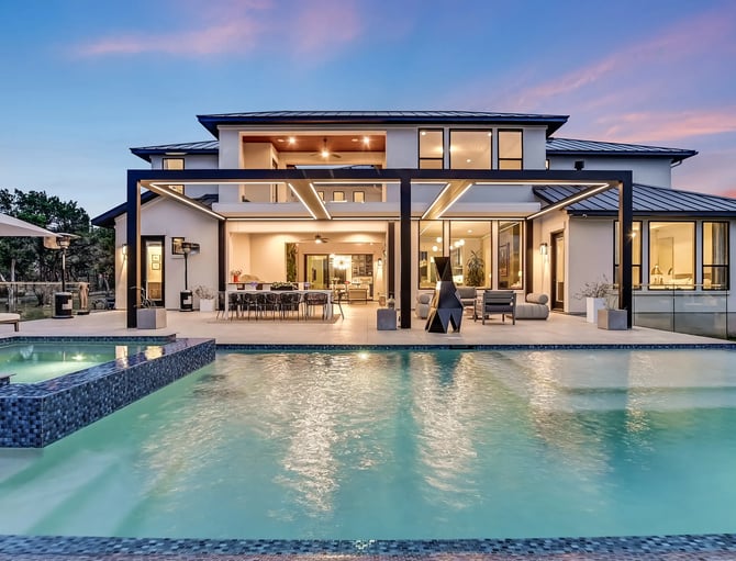 Modern two-story house with pool and spa at dusk, featuring glass walls, black metal pergolas, and lit interior spaces
